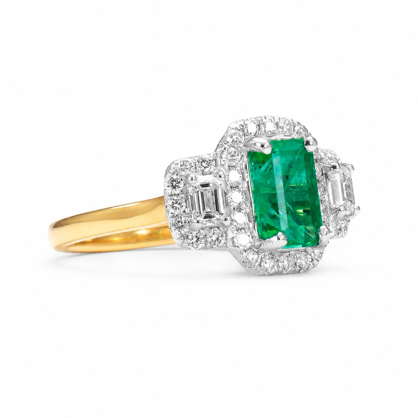 side view of a yellow gold ring with a large elongated emerald-cut Colombian emerald center stone  flanked by square cut diamonds and surrounding halo of diamonds, set against a clean white background.
