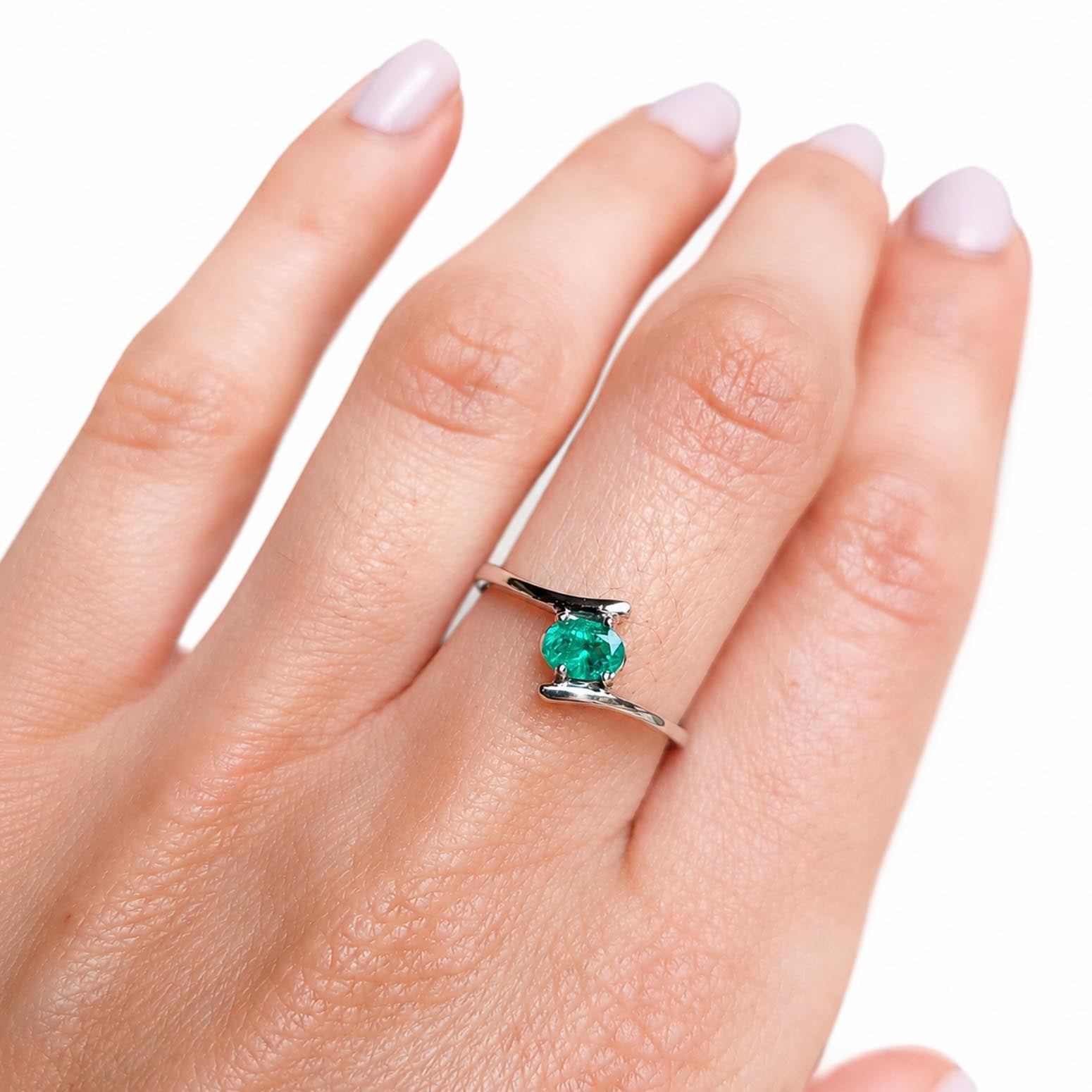 A white gold ring with an oval-cut Colombian emerald gemstone, shown worn on a hand with pale pink manicured nails against a white background.