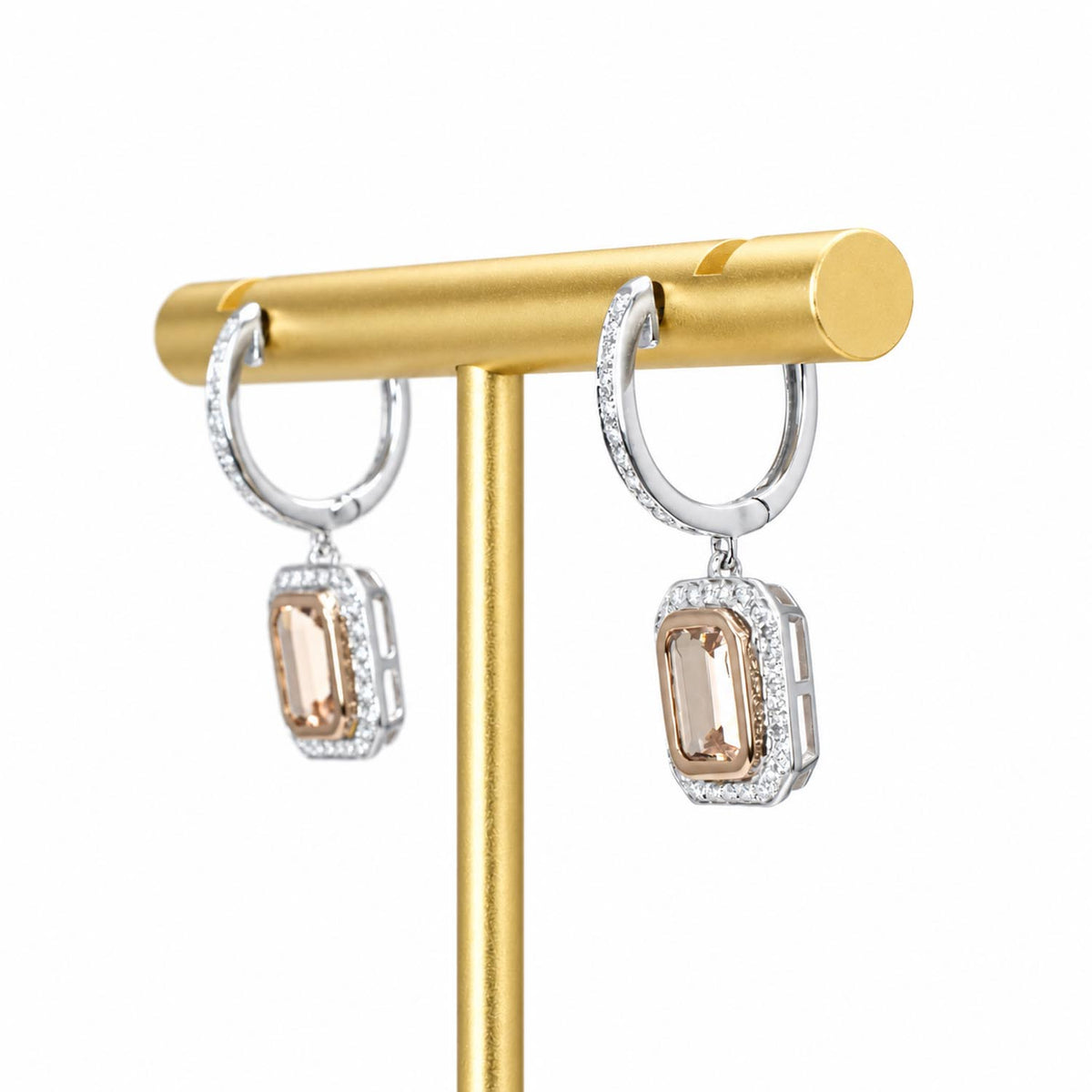 A pair of white gold hoop earrings with rectangular peach-colored morganites surrounded by diamonds, displayed on a gold stand against a white background.