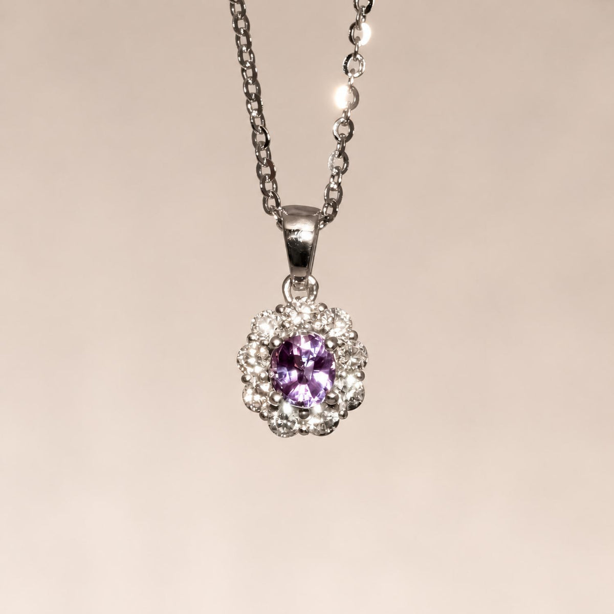 A platinum pendant necklace featuring a central alexandrite displaying its purple colour under incandescent light surrounded by a halo of small diamonds, suspended from a delicate silver chain against a soft beige background.