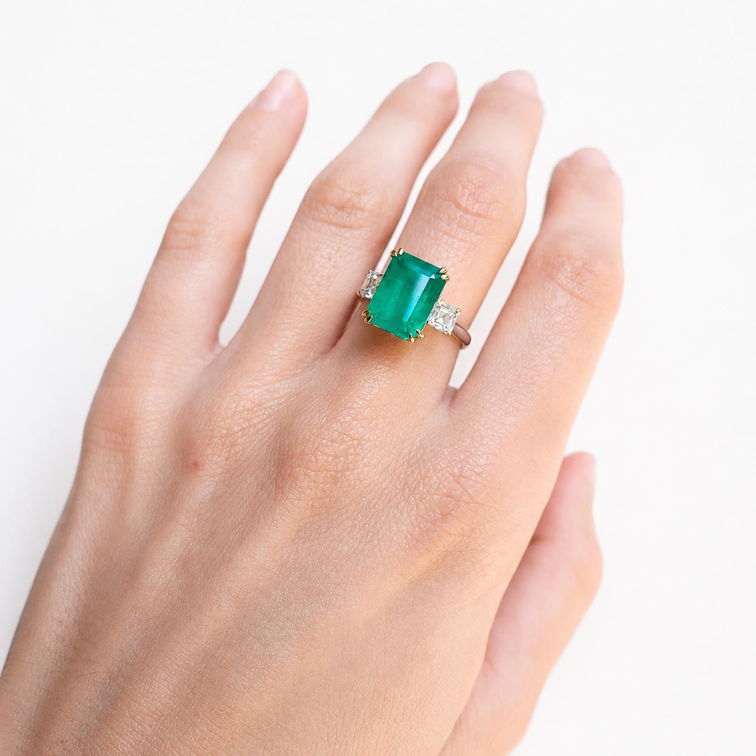 A close-up of a hand wearing a ring with an elongated Colombian emerald-cut center stone and side emerald cut square diamonds on a white background.