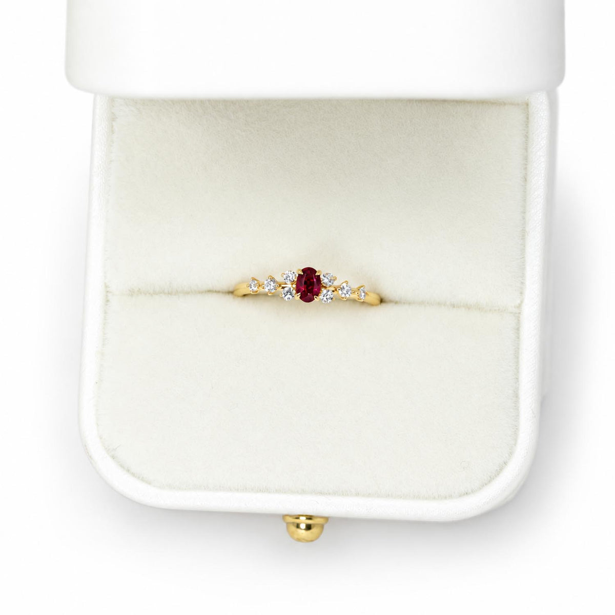A yellow gold ring with a central oval ruby and small diamonds side stones, displayed in an open white ring box on a white background.