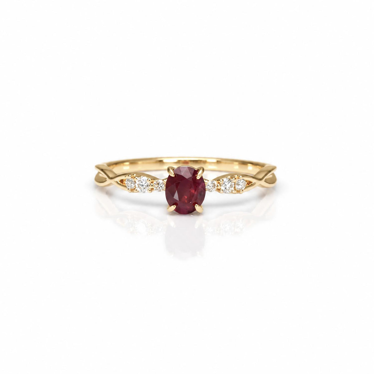 A yellow gold ring featuring an oval-cut red ruby center stone, flanked by small diamonds on a white background with a soft reflection.