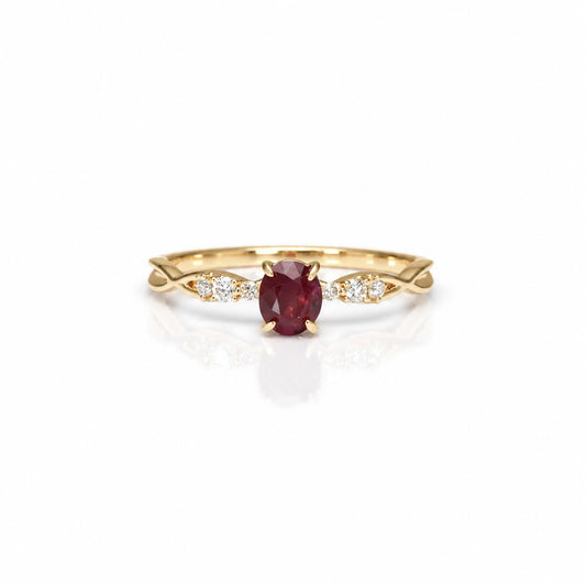 A yellow gold ring featuring an oval-cut red ruby center stone, flanked by small diamonds on a white background with a soft reflection.