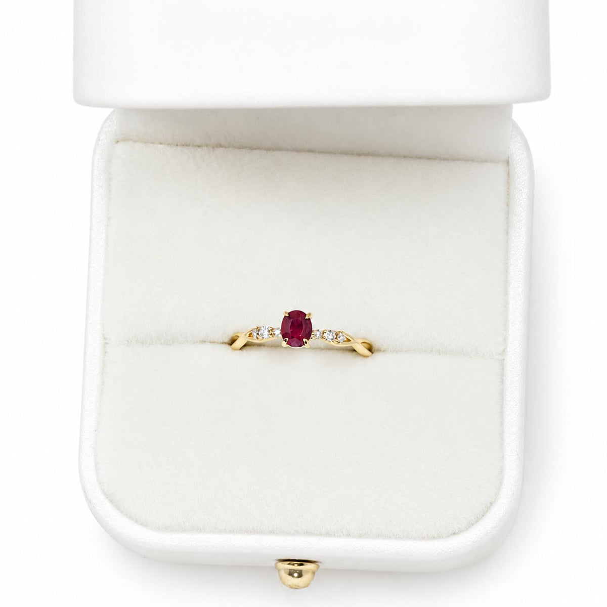 A yellow gold ring with an oval-cut red ruby center stone and small diamond accent stones on either side, displayed inside an open white ring box.