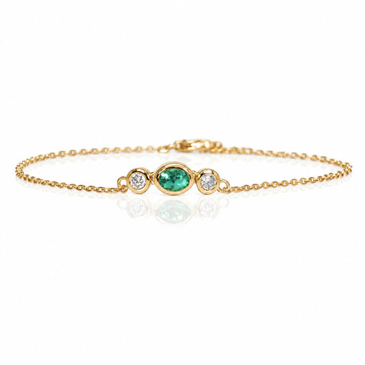 A delicate yellow gold chain bracelet featuring a central oval-cut Colombian emerald flanked by two small round diamonds in gold bezels, displayed on a clean white background with a soft reflection.