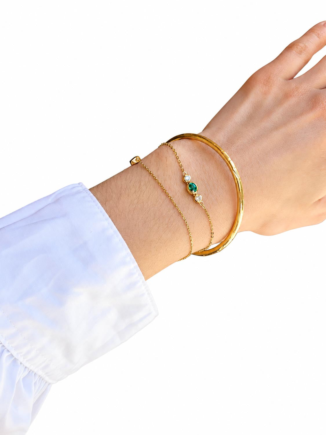 A close-up of a person's wrist wearing three delicate gold bracelets: a thick bangle, a fine chain with a small Colombian emerald stone and white diamond accents, and a thin chain with a small gold bead.