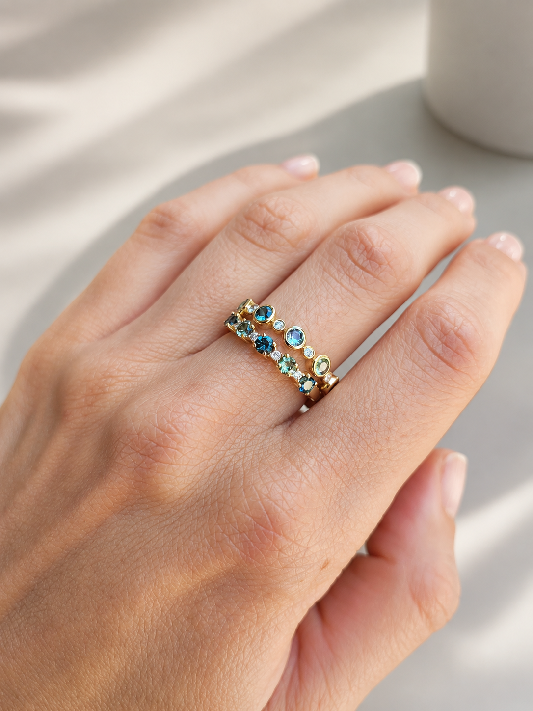 A close-up of a hand wearing two delicate gold bands set with Australian teal sapphires and accent diamonds photographed against a softly lit neutral background. 