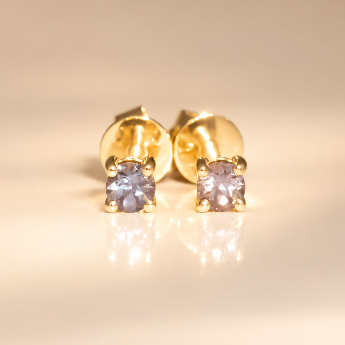 view under incandescent light of a pair of 18K yellow gold stud earrings with natural alexandrites as centre stones shown in a neutral background with a soft reflection