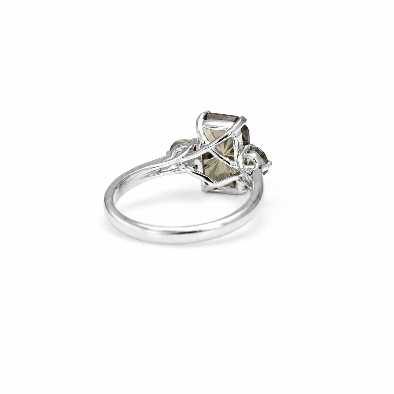 A white gold ring featuring a central rectangular clear gemstone wrapped in twisted metal, flanked by two small round teal green Australian sapphires, shown on a plain white background.