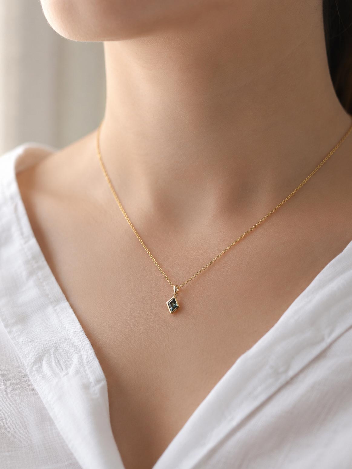 A close-up of a model wearing a delicate gold chain necklace with a small geometric diamond-shaped pendant featuring a natural unheated teal sapphire, styled with a white top.