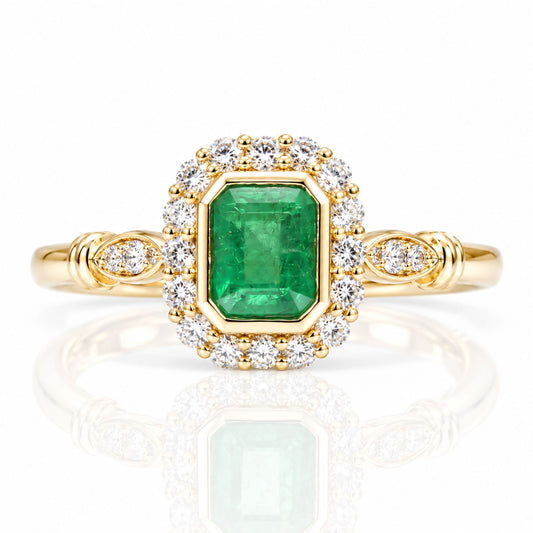 A yellow gold ring featuring a large emerald-cut Colombian emerald in a halo setting surrounded by small round diamonds, with additional diamonds on the band, shown on a reflective white background