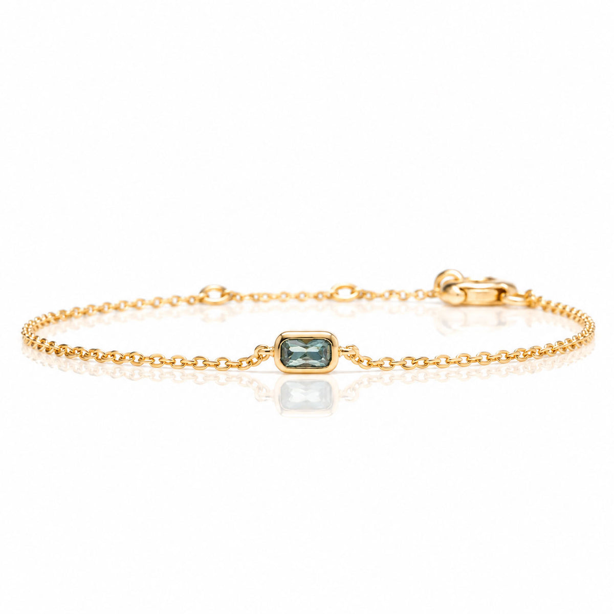 A delicate gold chain bracelet featuring a central rectangular bezel-set light blue sapphire, displayed on a clean white reflective surface.