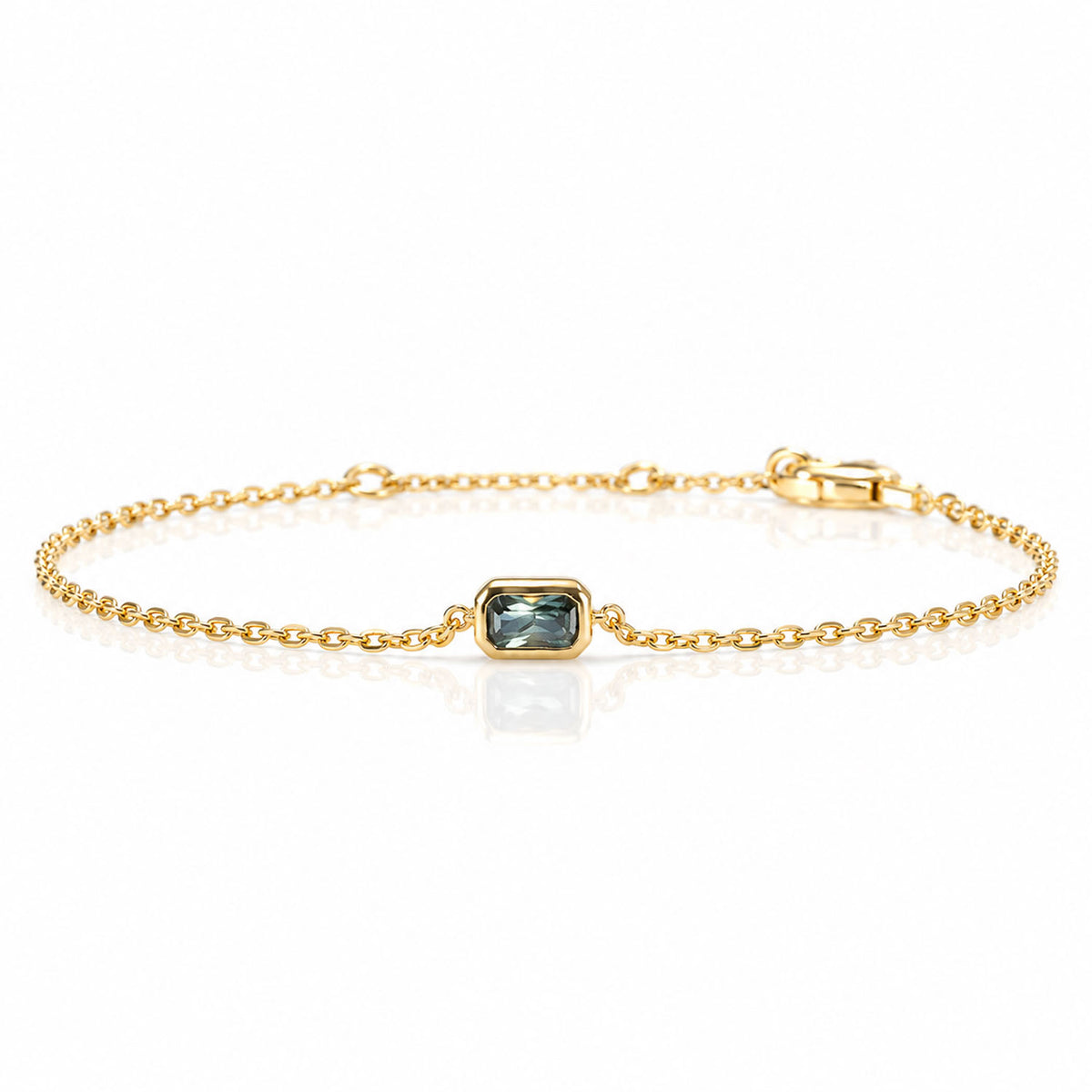 A delicate gold chain bracelet featuring a central rectangular bezel-set teal sapphire, displayed on a clean white background with a soft reflection.