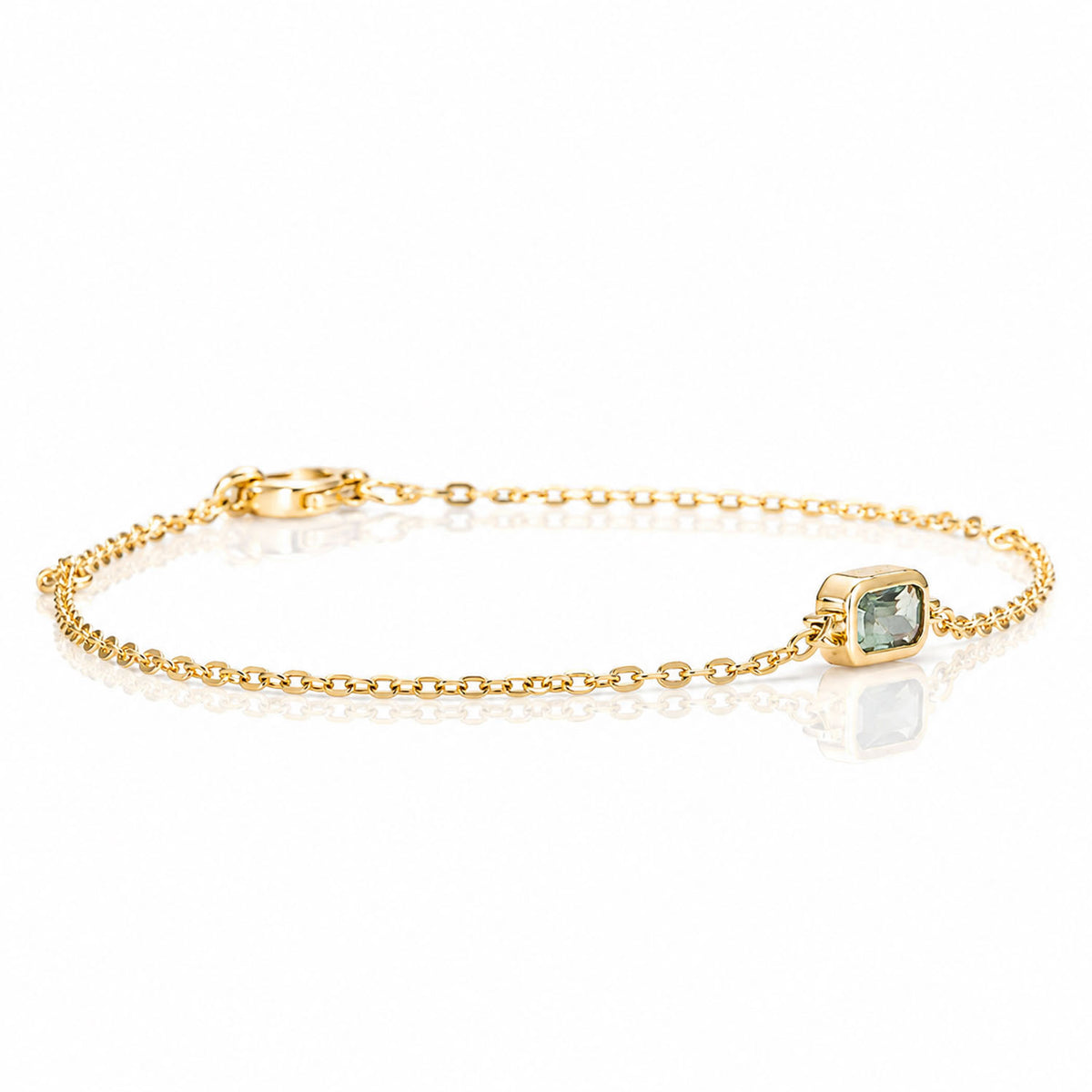 A delicate yellow gold chain bracelet with a small bezel-set light sapphire, displayed on a clean white reflective surface.
