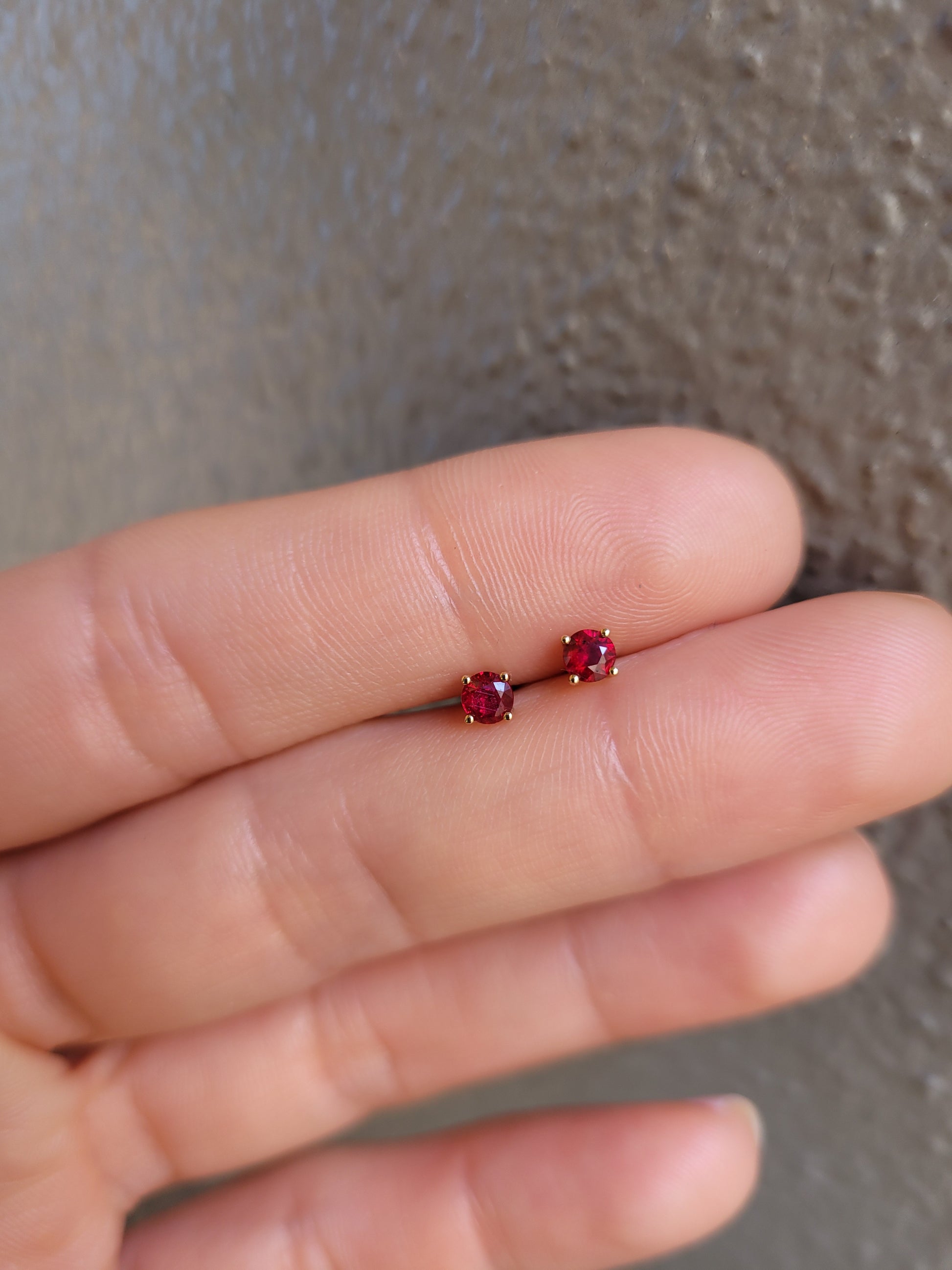 Gold stud earrings with ruby gemstones on a person's finger against a neutral background