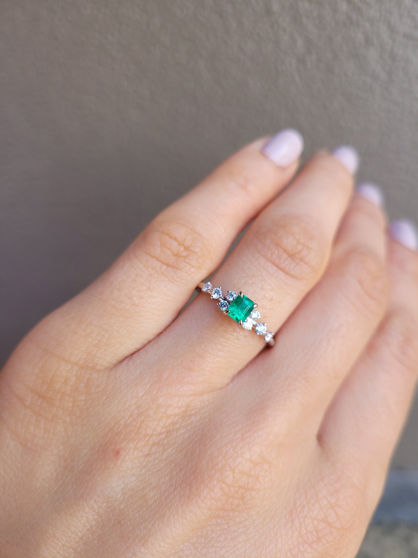 photo of platinum ring with emerald and diamonds on a hand in gray background