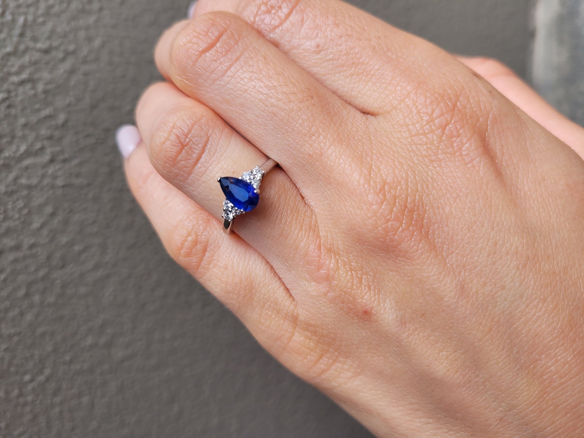 photo on hand of Platinum ring with a blue teardrop-shaped sapphire and smaller diamonds on a light gray background