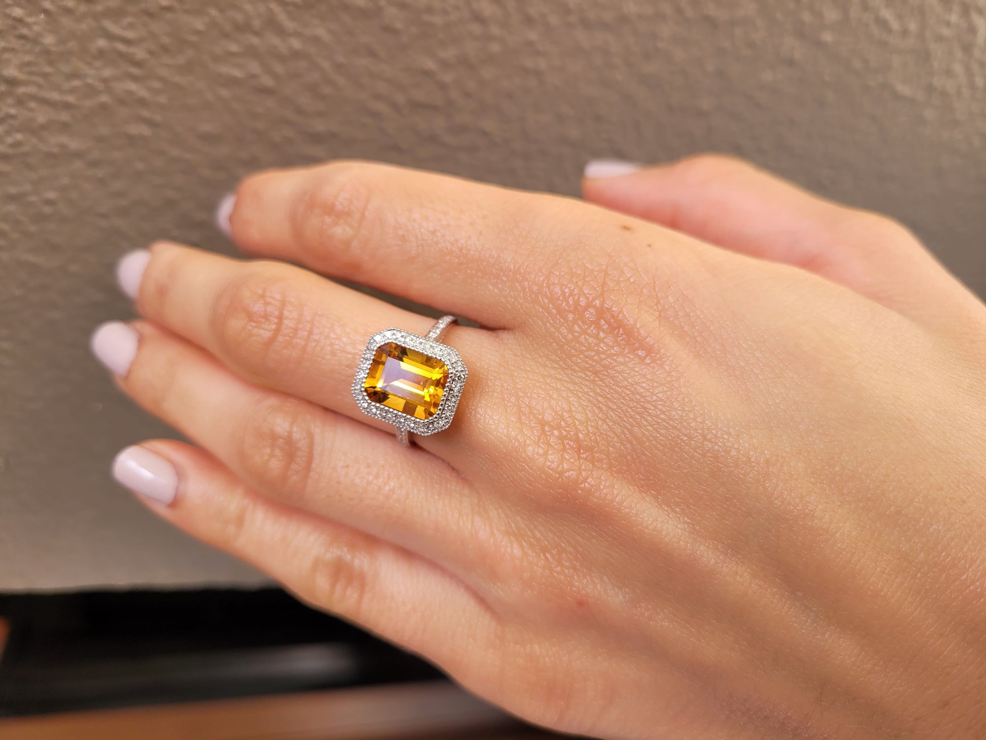 photo on hand of Citrine and diamond halo ring in 18K white gold in light gray background