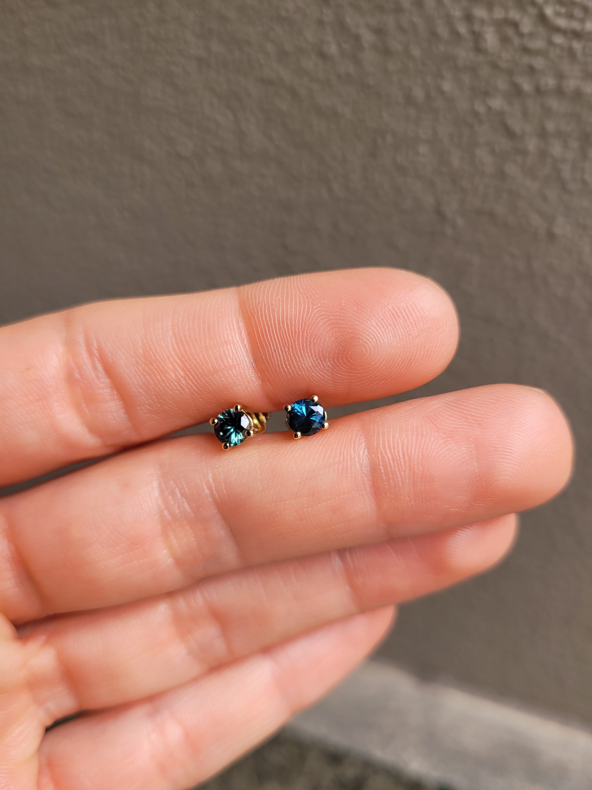 photo of hand holding a pair of round natural Australian teal sapphire studs in neutral background