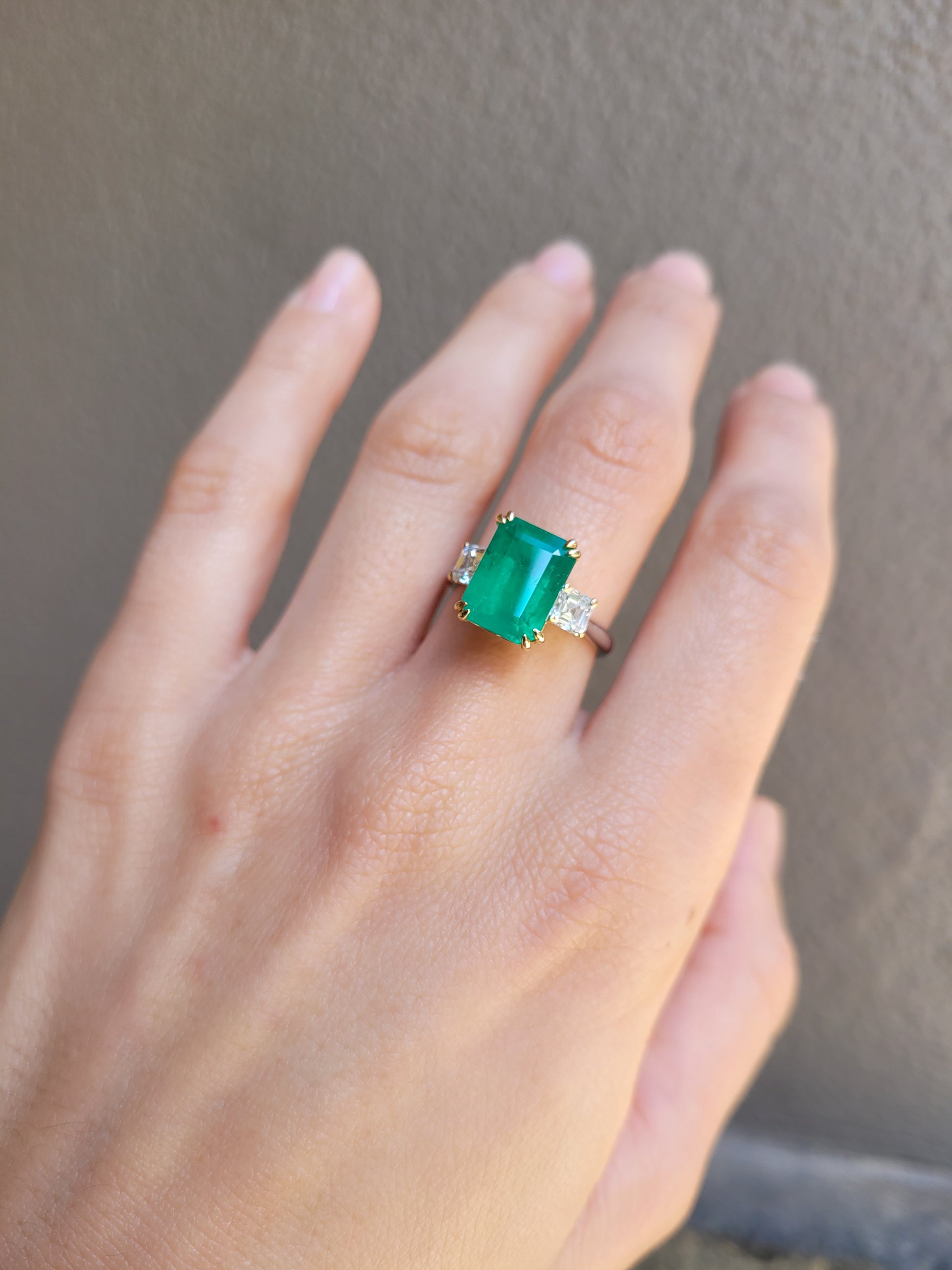 Hand wearing a Colombian Emerald and Diamond Ring in 18K Yellow Gold – East West Emerald Cut in neutral background