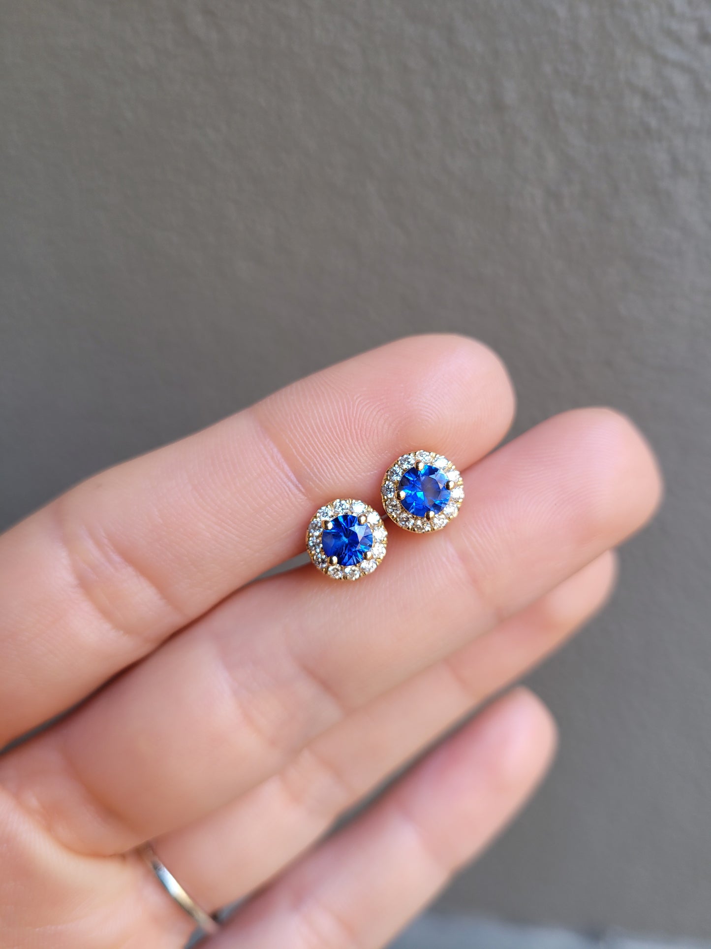 photo of Blue Sapphire and Diamond Halo Stud Earrings in 18K Yellow Gold held between fingers in neutral background