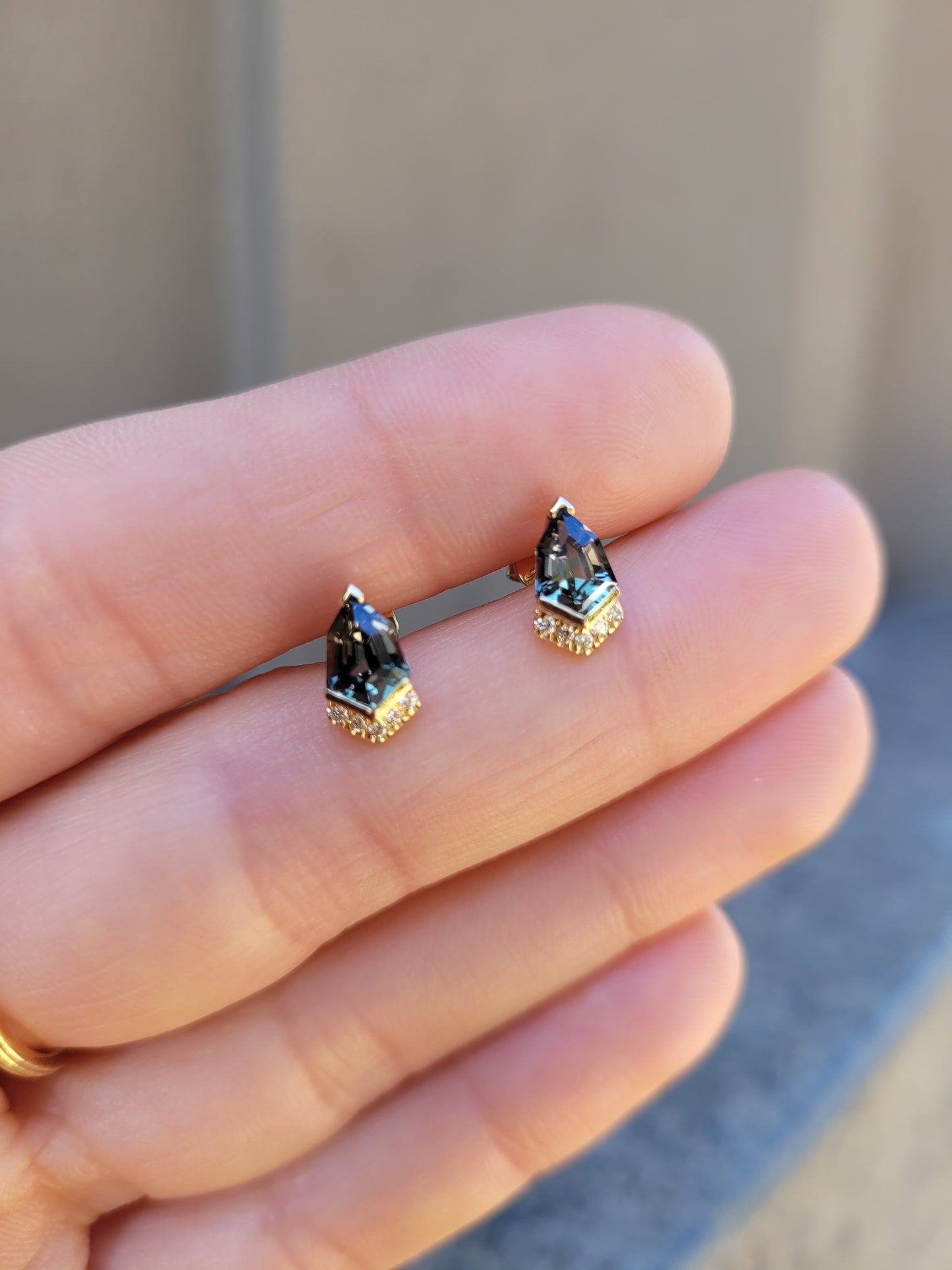 Shield Cut Teal Sapphire Diamond Stud Earrings in 18K Yellow Gold held in between fingers