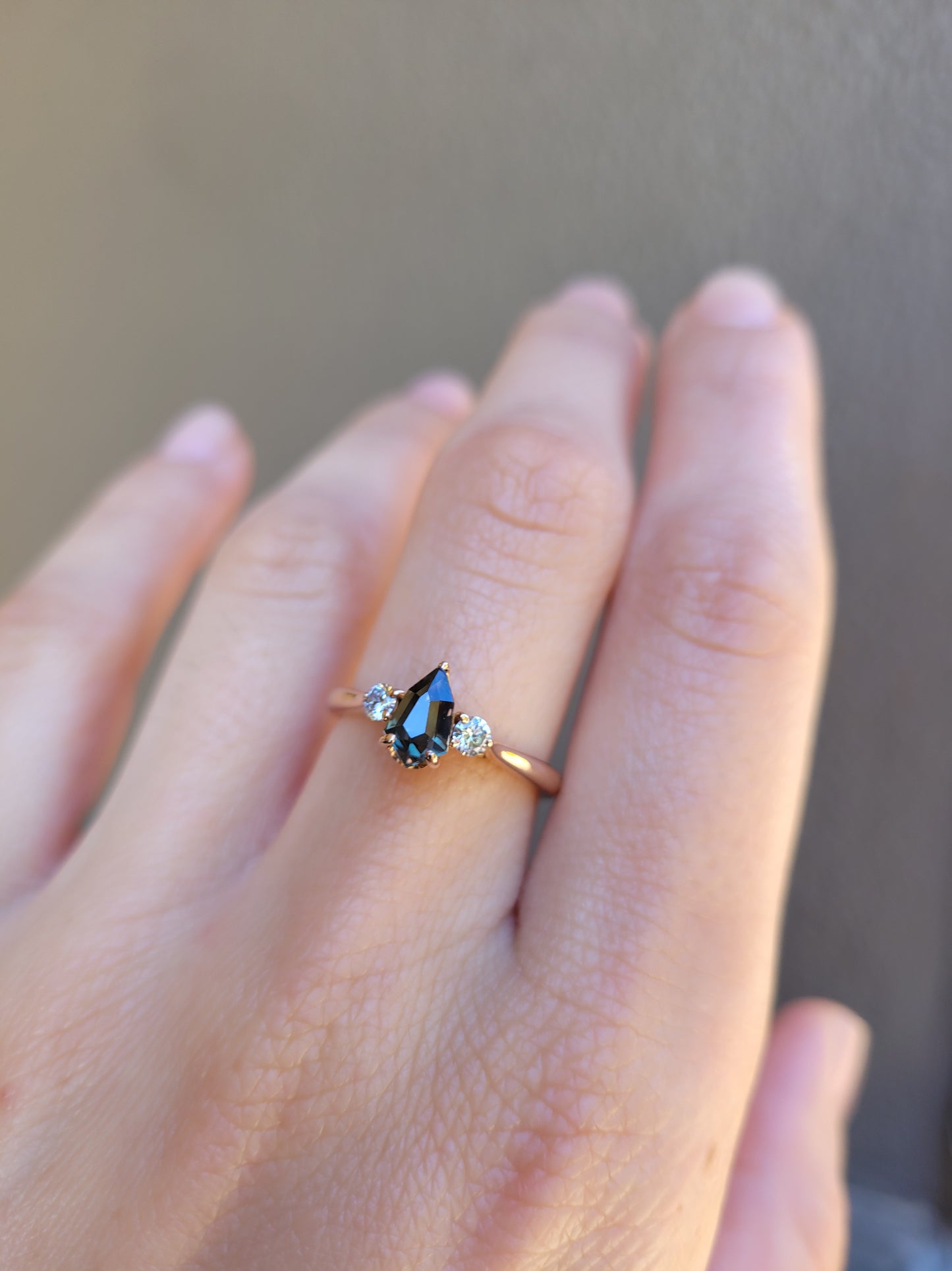 photo on hand of Shield Cut Teal Sapphire Diamond Ring in 18K Rose Gold on neutral background