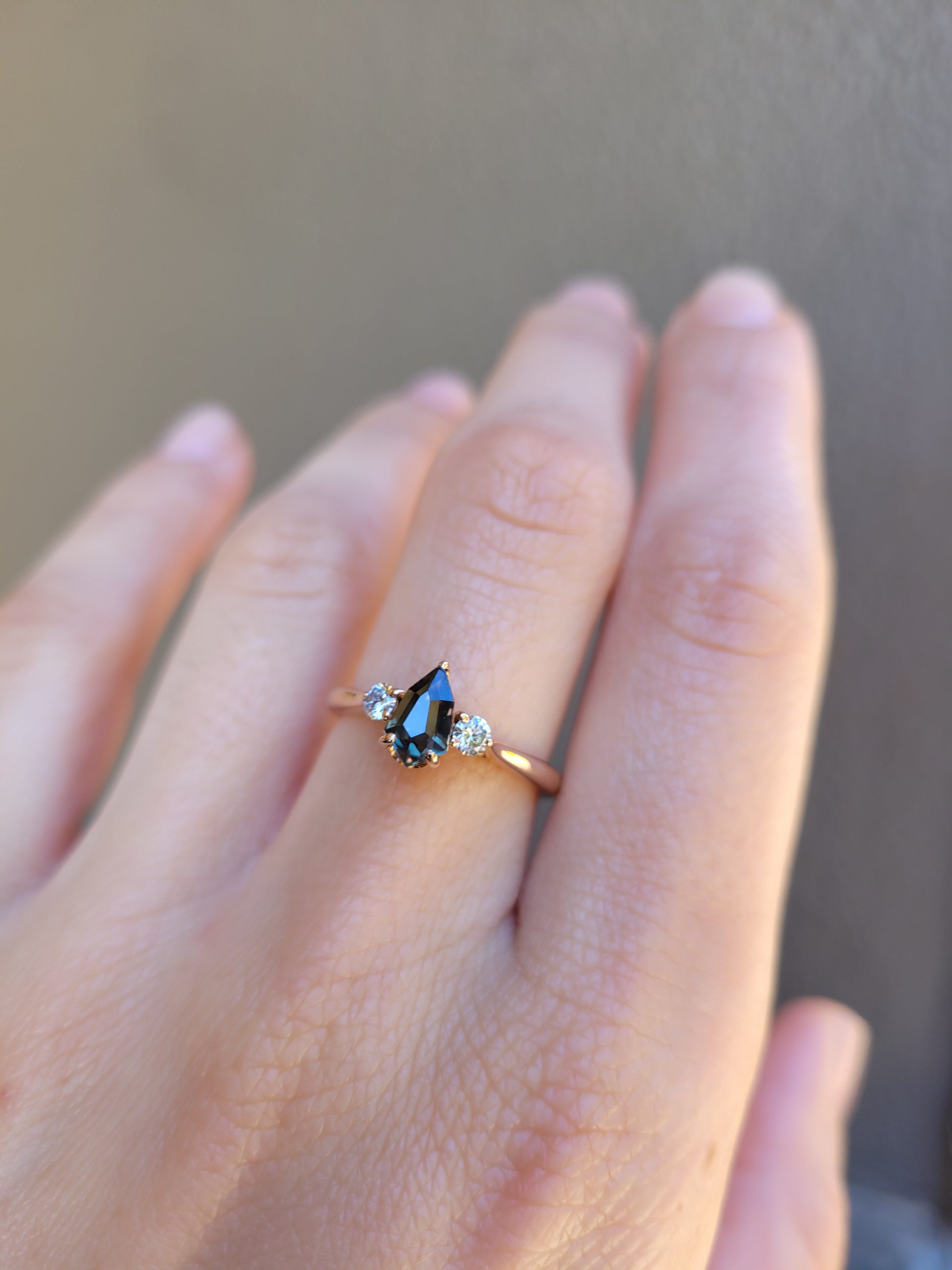 photo on hand of Shield Cut Teal Sapphire Diamond Ring in 18K Rose Gold on neutral background