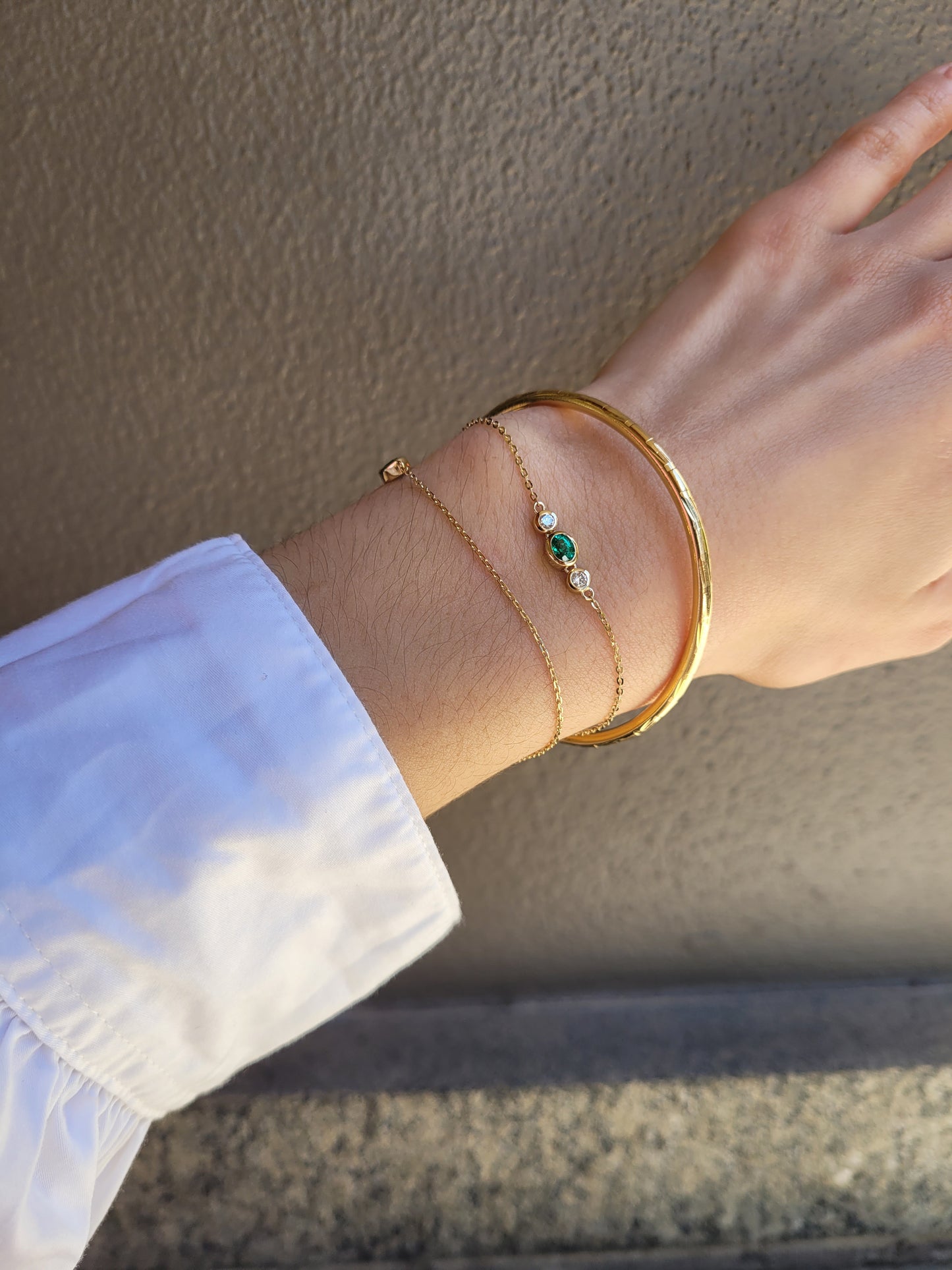 A close-up of a person's wrist wearing three delicate gold bracelets, including a Colombian emerald and diamonds dainty bracelet set against a textured beige wall.