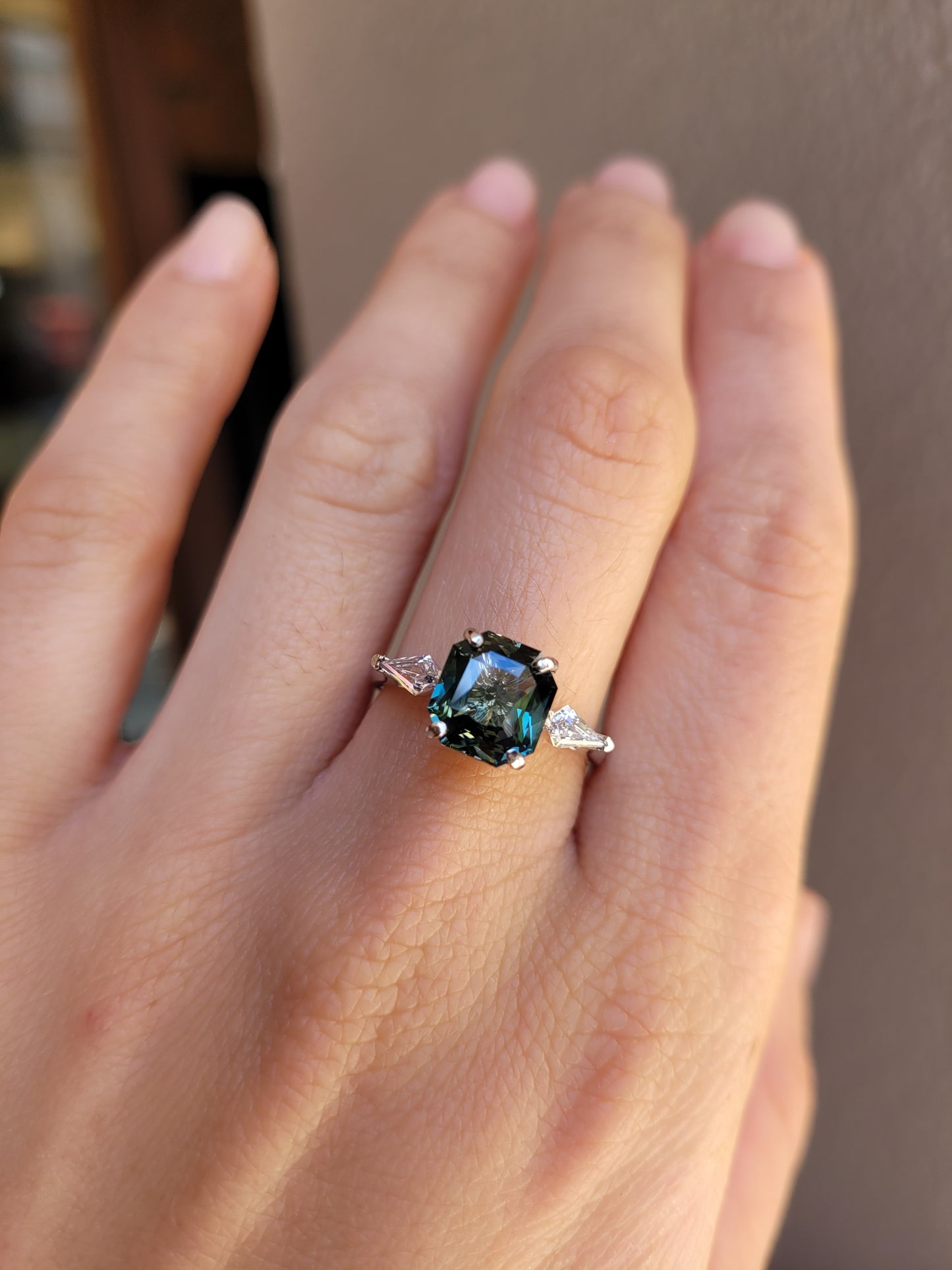 A close-up of a hand wearing a Natural Teal Sapphire & Kite Diamond Ring in Platinum on a warm background.