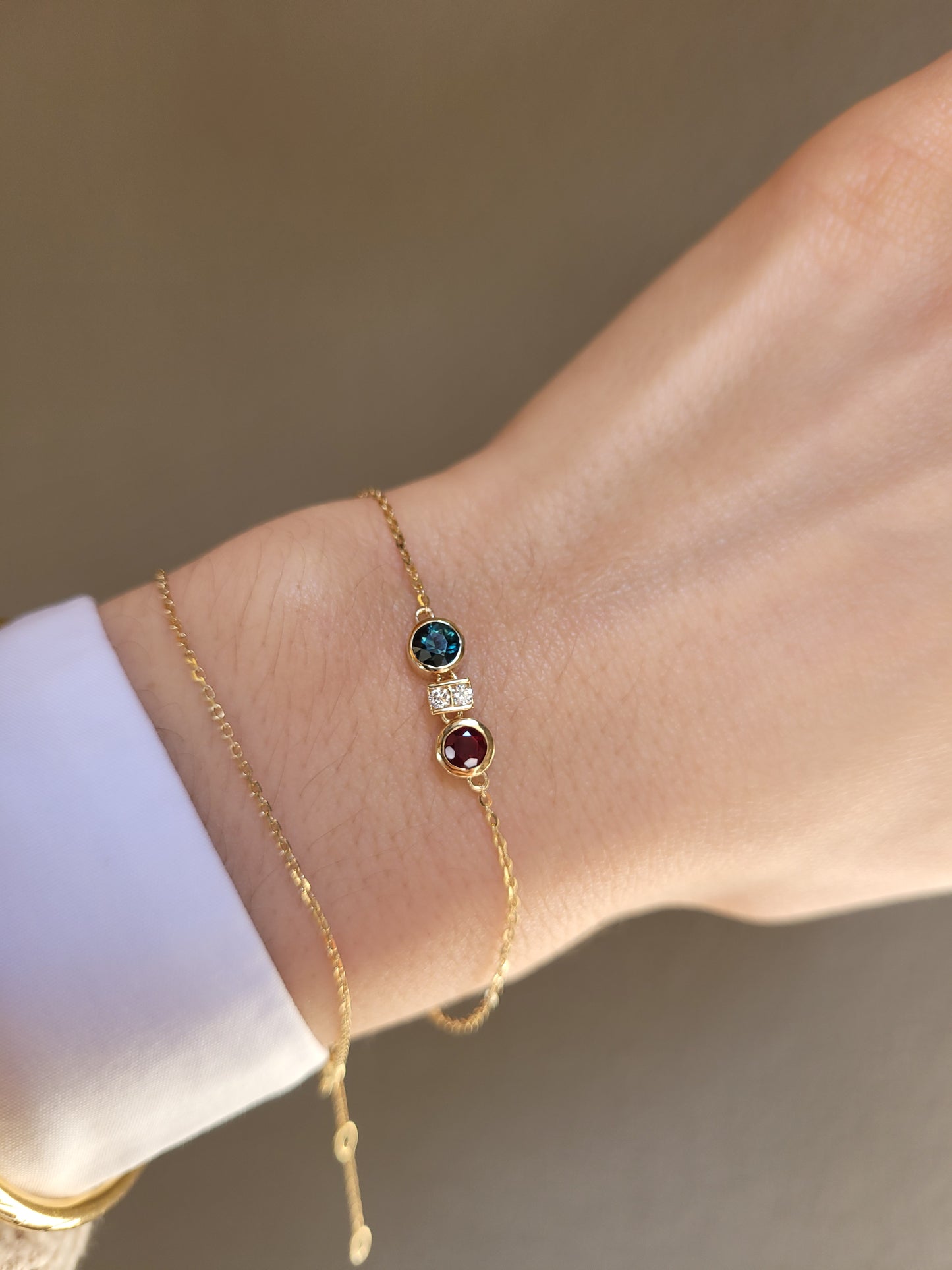 a close-up photo of a wrist wearing a delicate gold chain bracelet featuring two round bezel-set gemstones, one natural ruby and one Australian teal sapphire, separated by accent diamonds shown on a white 
