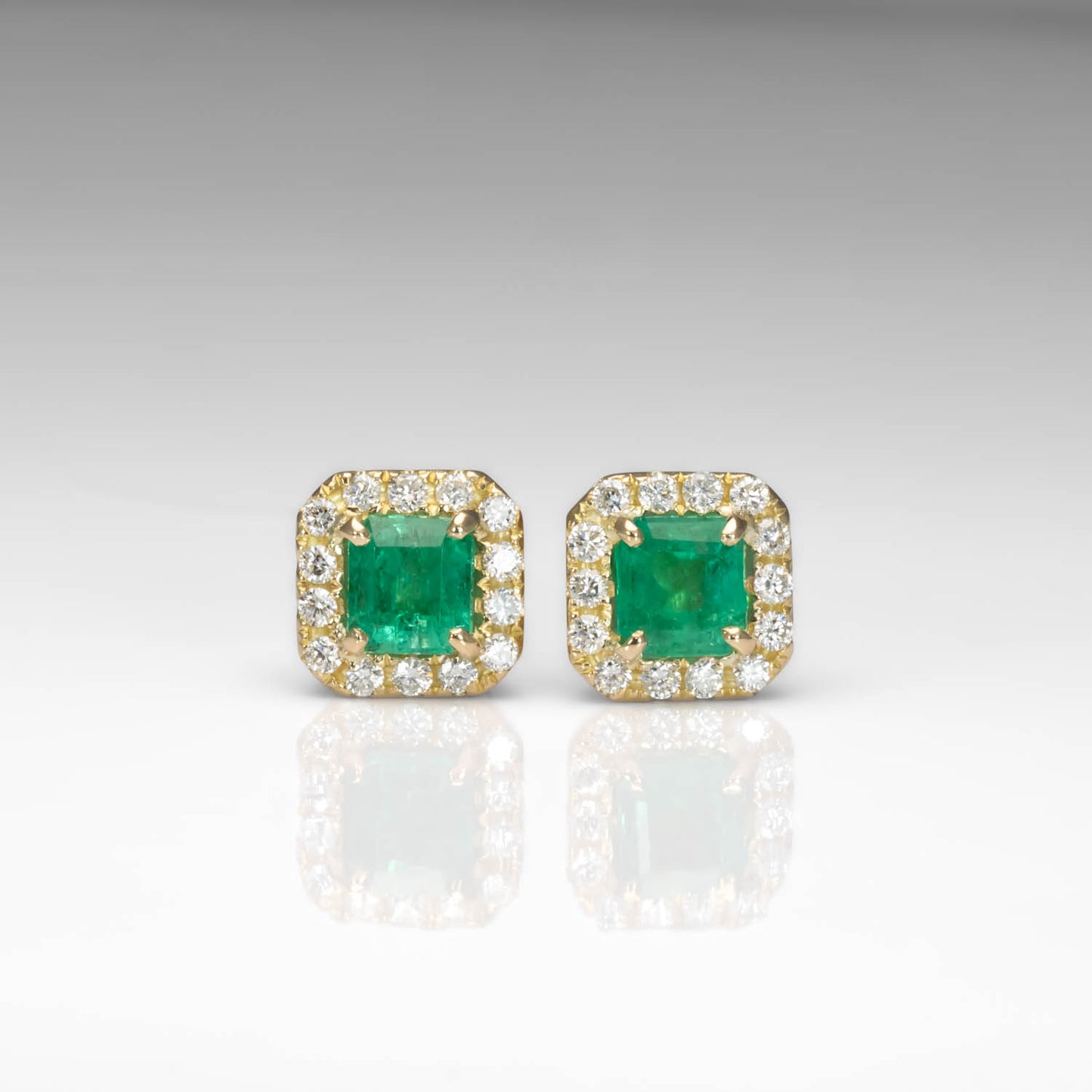 A pair of gold square stud earrings featuring green emerald-cut center stones surrounded by a halo of small clear stones, shown on a light gray reflective surface.