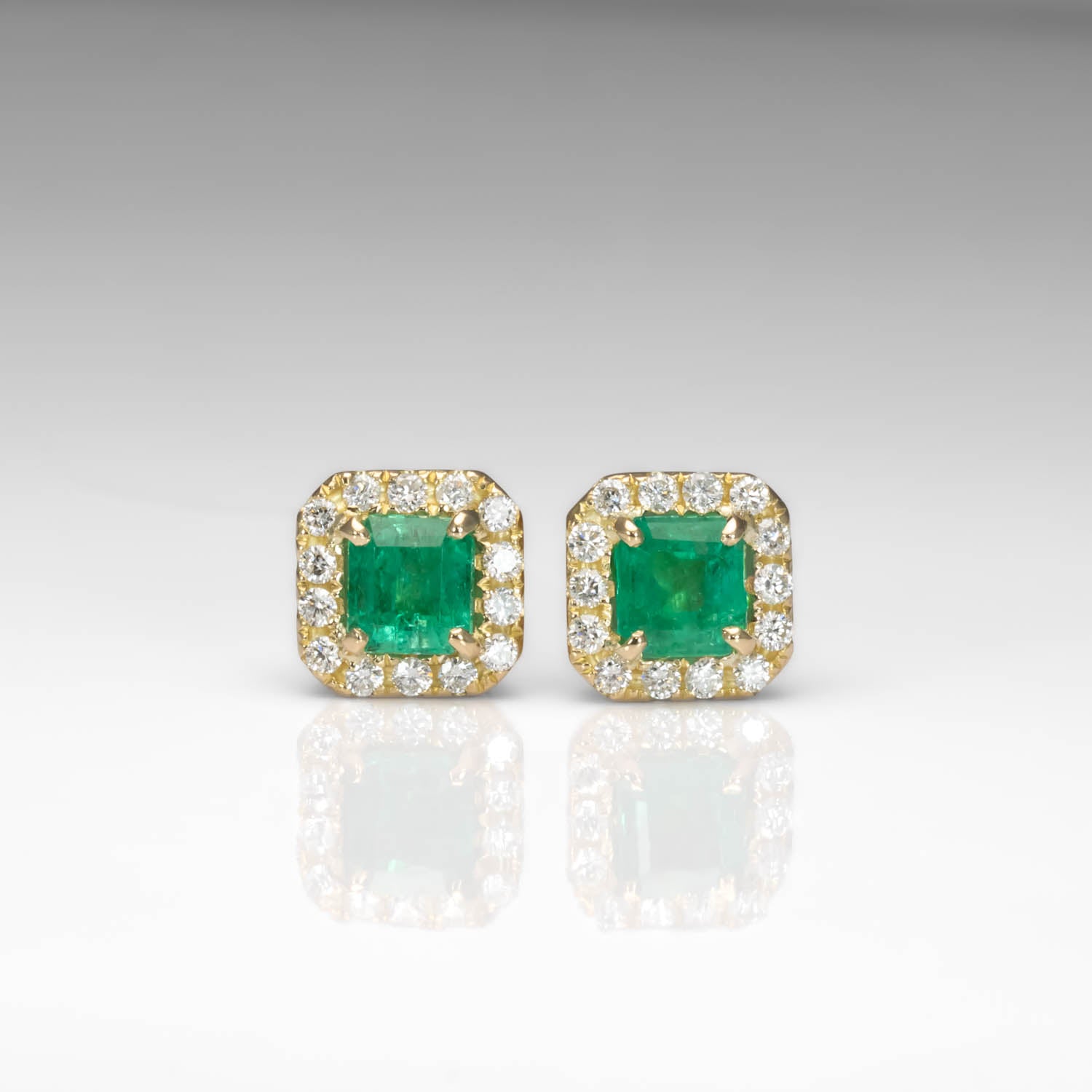 A pair of gold square stud earrings featuring green emerald-cut center stones surrounded by a halo of small clear stones, shown on a light gray reflective surface.