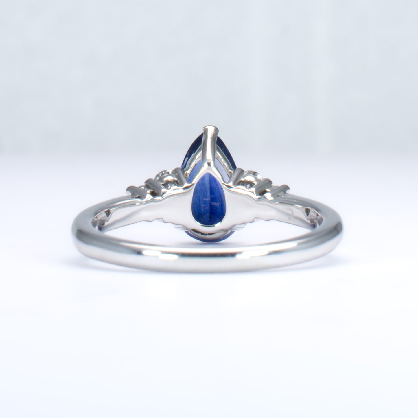 back view of Platinum ring with a blue teardrop-shaped sapphire and smaller diamonds on a light gray background
