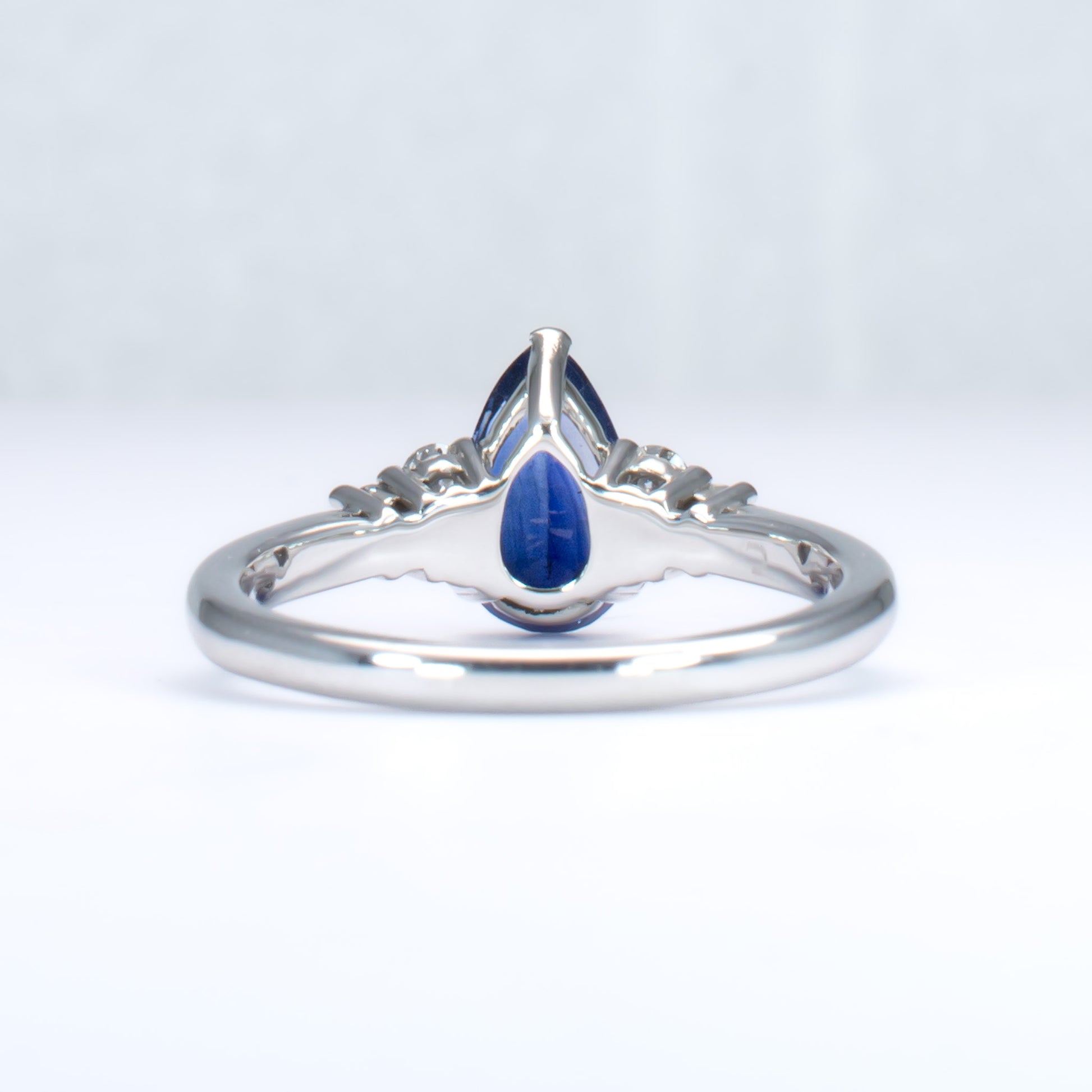 back view of Platinum ring with a blue teardrop-shaped sapphire and smaller diamonds on a light gray background
