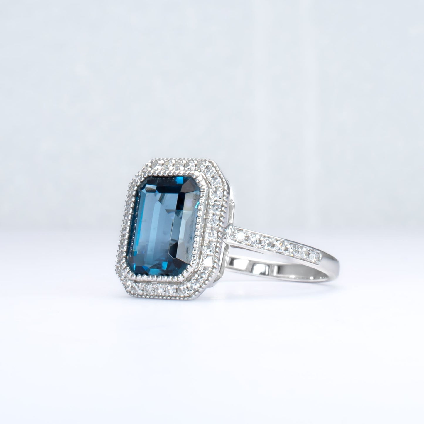 side view of White gold ring with London blue topaz and diamond halo in light gray background