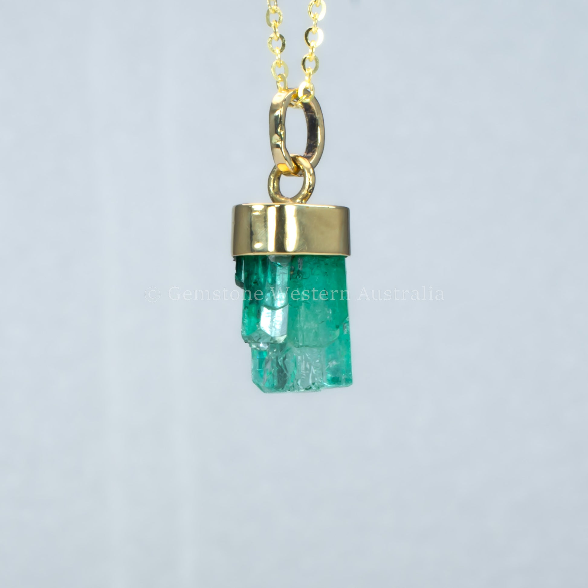 second perspective of Gold pendant with a rough emerald crystal on a light gray background