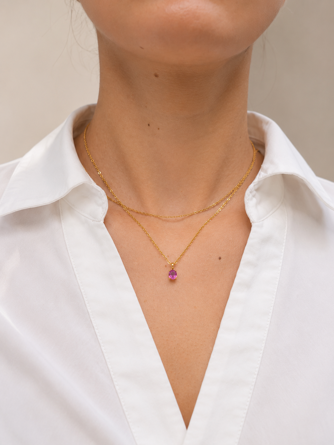 A close-up of a model wearing a delicate gold necklace with a small pink sapphire pendant, styled with a white collared shirt.