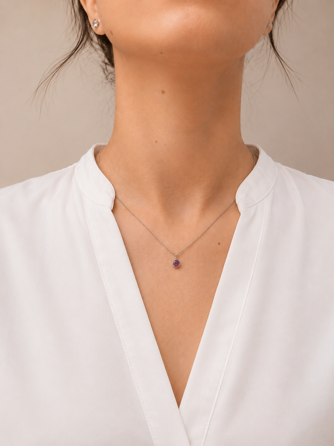 A close-up of a model wearing a delicate silver chain necklace with a small purple pink sapphire pendant, paired with a white V-neck blouse.