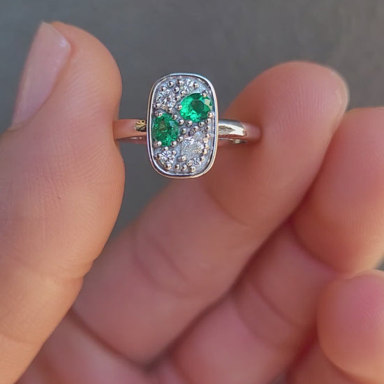 video of a Natural Colombian Emerald & Diamond Composition Ring in Platinum held between two fingers shown against a textured gray background.