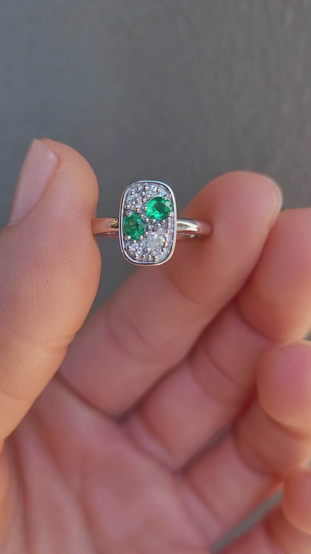 video of a Natural Colombian Emerald & Diamond Composition Ring in Platinum held between two fingers shown against a textured gray background.