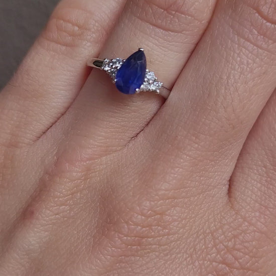 video on hand of Platinum ring with a blue teardrop-shaped sapphire and smaller diamonds on a gray background