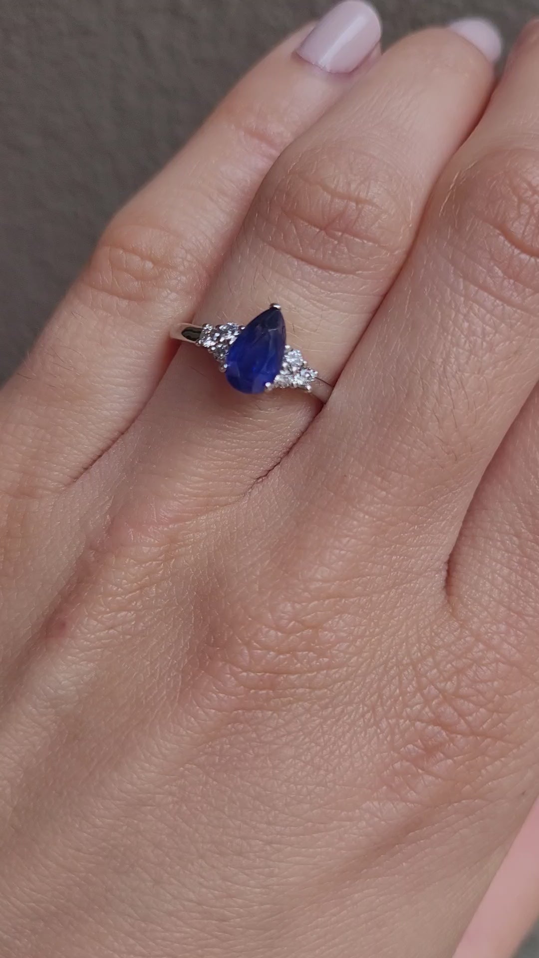 video on hand of Platinum ring with a blue teardrop-shaped sapphire and smaller diamonds on a gray background