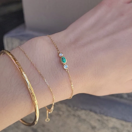 A close-up video of a person's wrist wearing three delicate gold bracelets, including a Colombian emerald and diamonds dainty bracelet set against a textured beige wall.