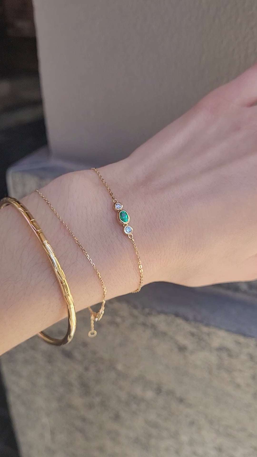 A close-up video of a person's wrist wearing three delicate gold bracelets, including a Colombian emerald and diamonds dainty bracelet set against a textured beige wall.