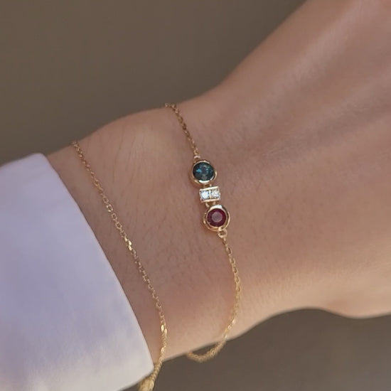 close-up video of a hand wearing a delicate gold chain bracelet featuring two round bezel-set gemstones, one natural ruby and one Australian teal sapphire, separated by accent diamonds shown on a neutral