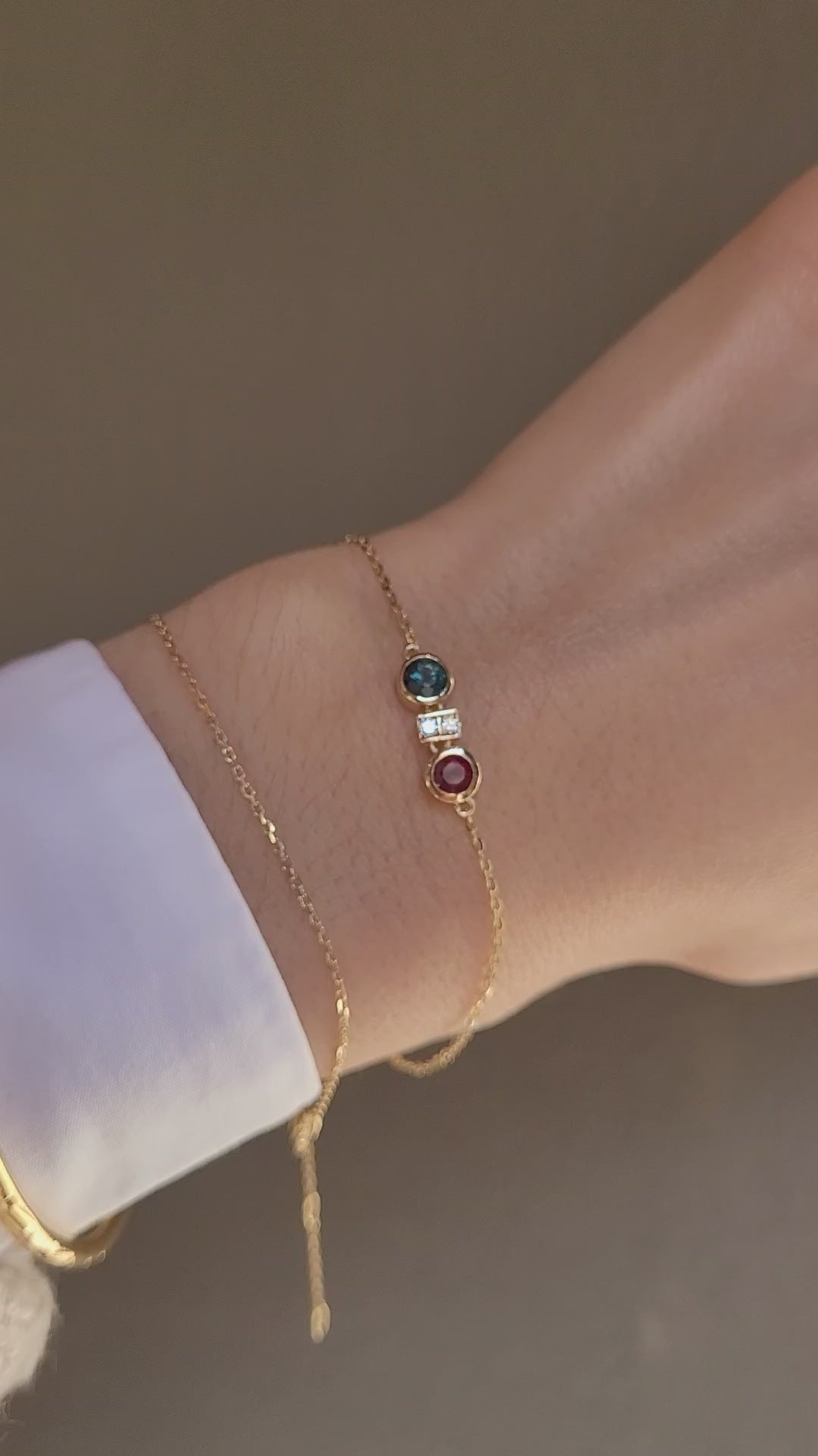 close-up video of a hand wearing a delicate gold chain bracelet featuring two round bezel-set gemstones, one natural ruby and one Australian teal sapphire, separated by accent diamonds shown on a neutral