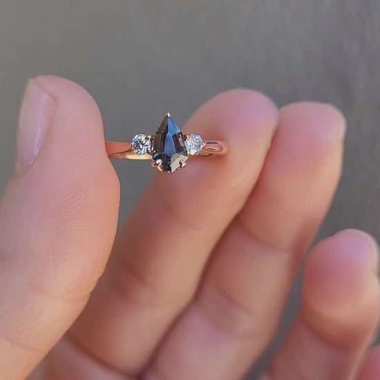 video of Shield Cut Teal Sapphire Diamond Ring in 18K Rose Gold in between fingers on neutral background