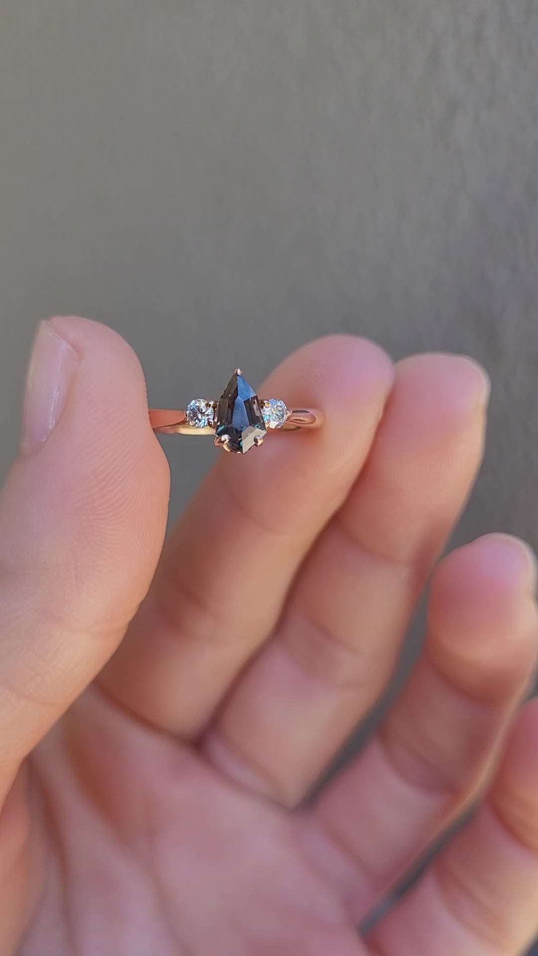 video of Shield Cut Teal Sapphire Diamond Ring in 18K Rose Gold in between fingers on neutral background